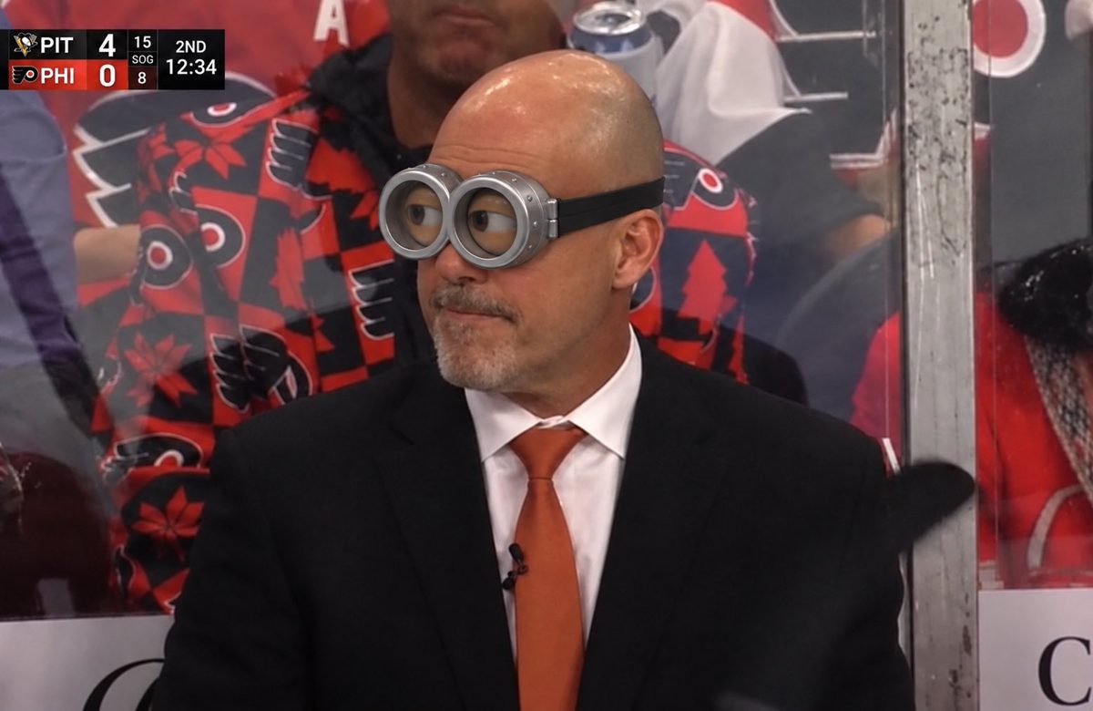The Flyers coach behind the bench wearing Minion goggles, with the scoreboard reading Pittsburgh 4, Philadelphia 0 and shots on goal 15 to 8 in the second period